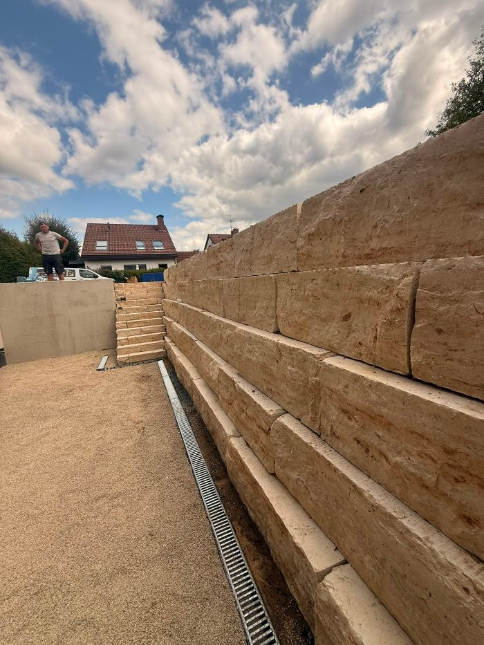 Hohe Blockstufenmauer aus Sandstein mit Entwässerungsrinne entlang der Kiesfläche